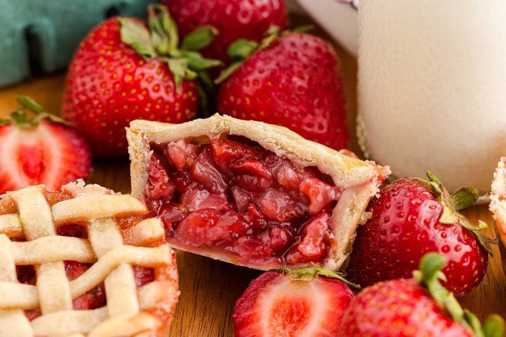 A sliced mini strawberry pie with a flaky crust, showing a juicy strawberry filling, surrounded by fresh whole and halved strawberries.