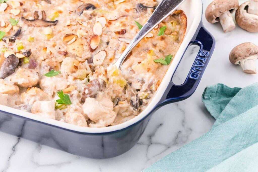 A baked casserole with chicken, mushrooms, sliced almonds, and herbs in a blue Staub dish, with a serving spoon and raw mushrooms nearby on a marble surface.