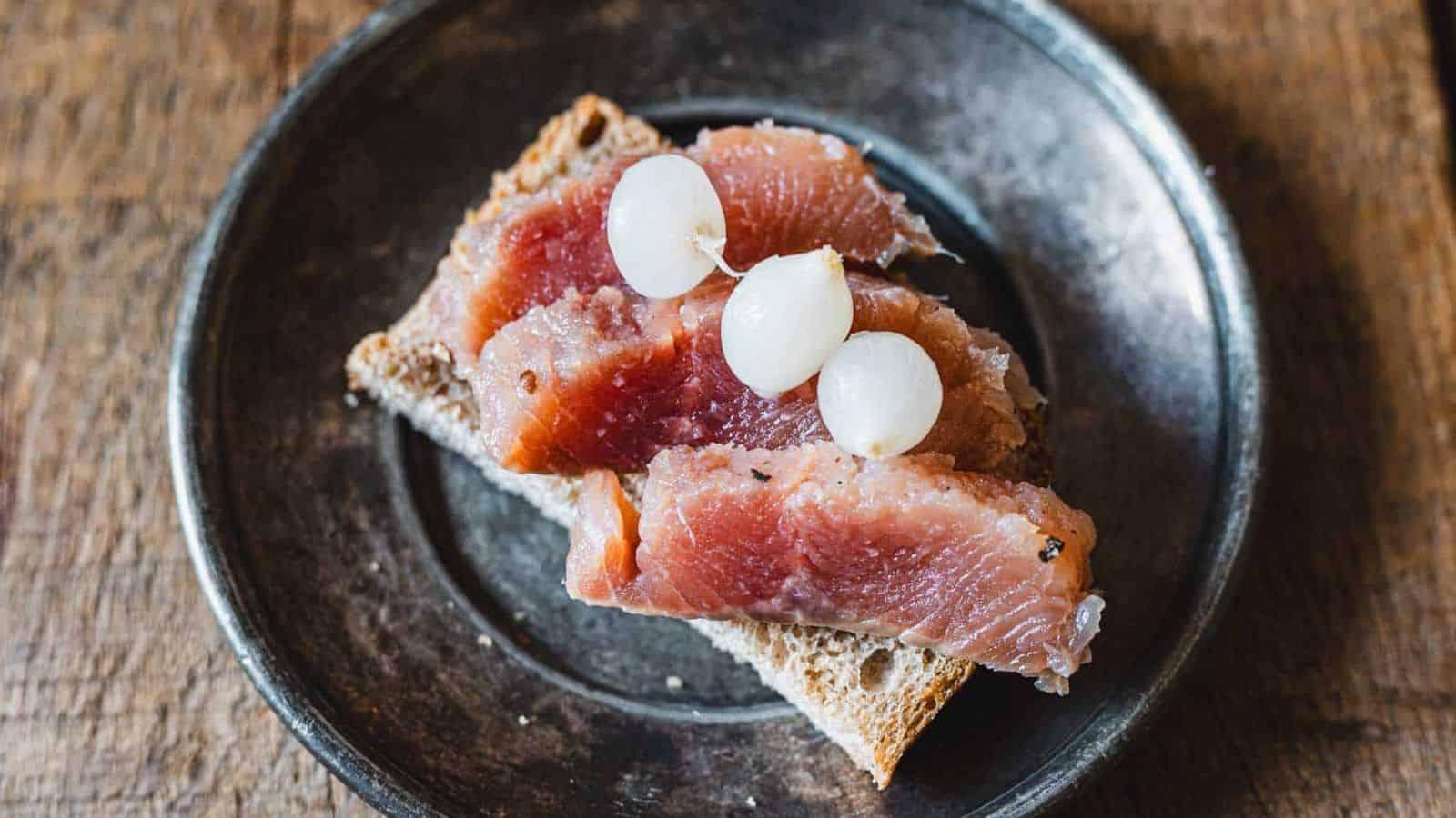 A slice of brown bread topped with three pieces of cured fish and three small white onions on a round, dark plate. The plate is placed on a wooden surface.