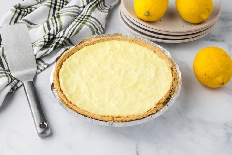 A lemon pie with a graham cracker crust sits on a marble surface next to whole lemons, stacked plates, a cloth napkin, and a metal pie server.