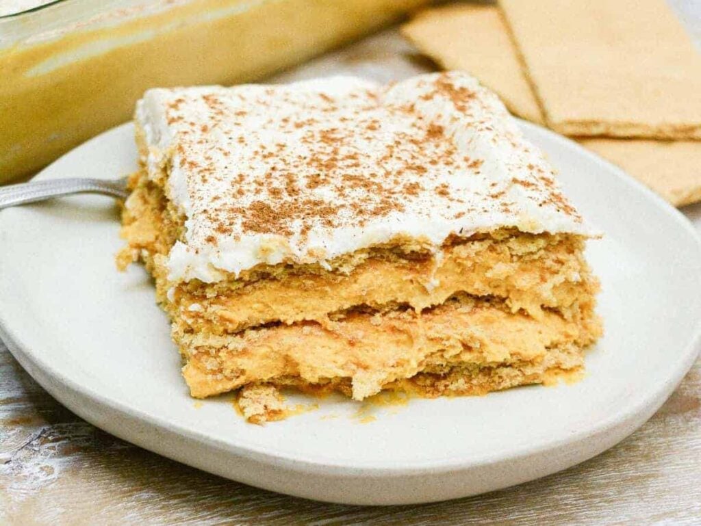A square slice of layered pumpkin dessert topped with whipped cream and cinnamon, served on a white plate with graham crackers in the background.