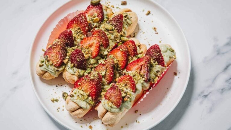 Three eclairs topped with green cream, sliced strawberries, and chopped pistachios are arranged on a white plate.