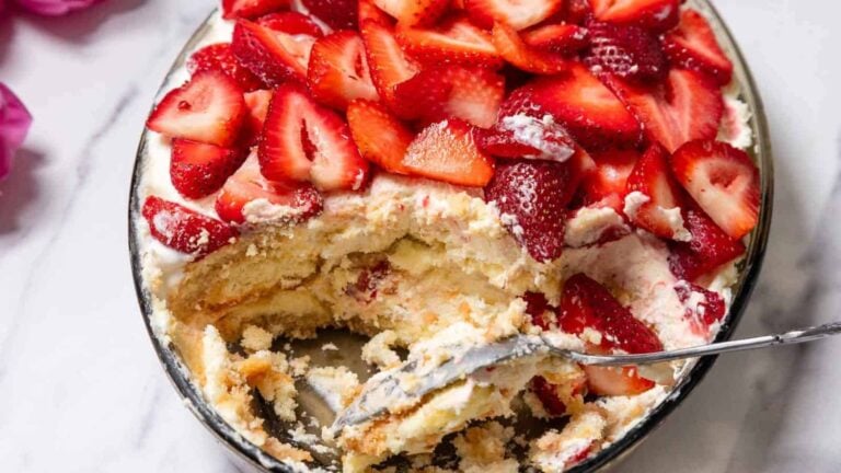 A glass dish filled with a layered dessert of whipped cream, sliced strawberries, and cake, with a serving spoon and a portion removed.