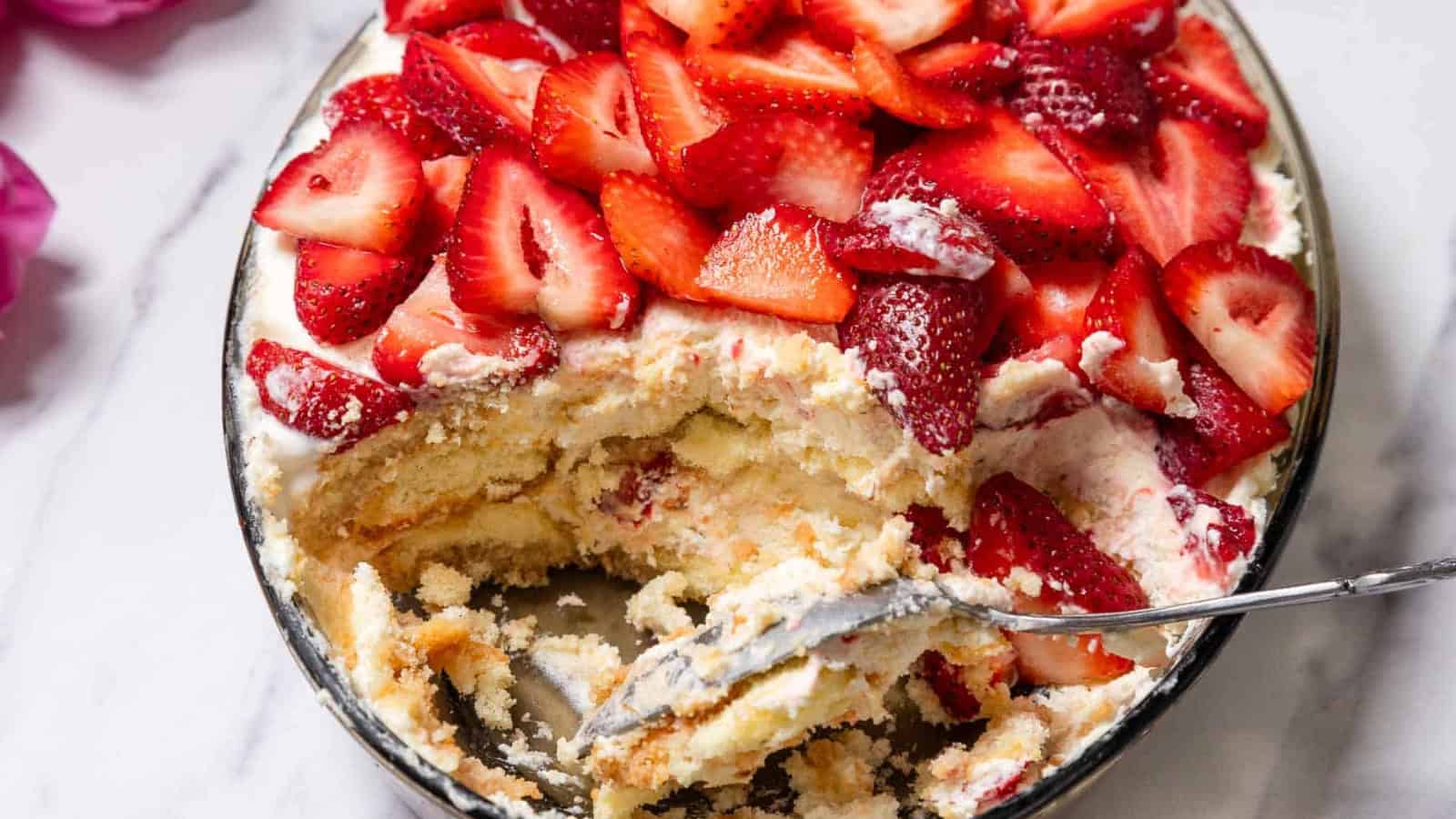 A dish of strawberry tiramisu with a spoon and a portion already served, topped with fresh sliced strawberries.