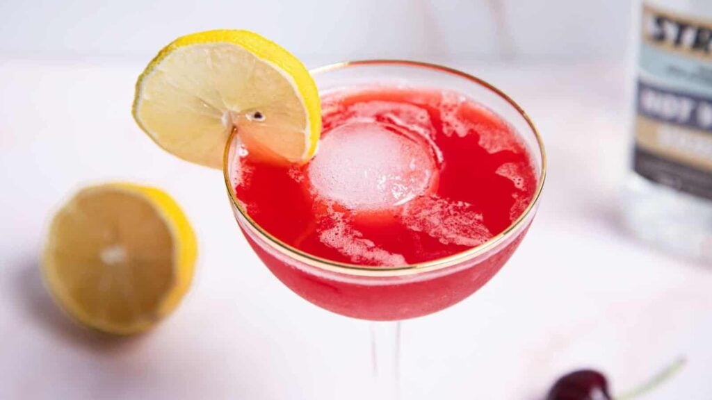 A red cocktail in a coupe glass with a lemon wheel garnish, a round ice cube, half a lemon, and a cherry nearby.