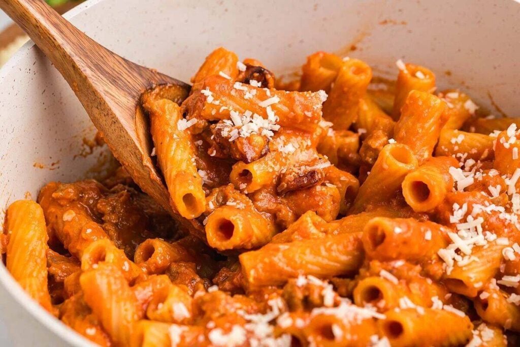 A close-up of rigatoni pasta mixed with tomato sauce, ground meat, and grated cheese, being stirred with a wooden spoon in a white pan.