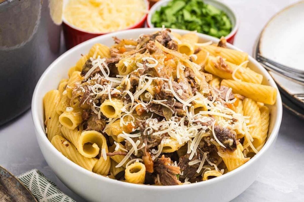 A bowl of rigatoni pasta topped with shredded meat and grated cheese, with bowls of shredded cheese and chopped herbs in the background.