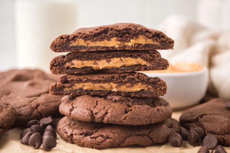 Stack of chocolate cookies with peanut butter filling, surrounded by more cookies and chocolate chips, with a bowl of peanut butter and a glass of milk in the background.