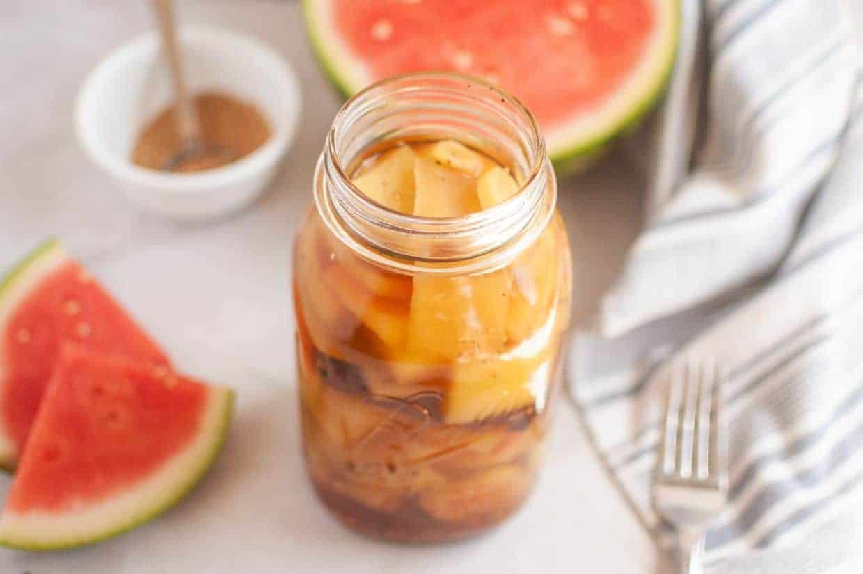 A jar of pickled watermelon rind sits on a white surface, with watermelon slices, a striped cloth, a fork, and a small bowl of spices in the background.