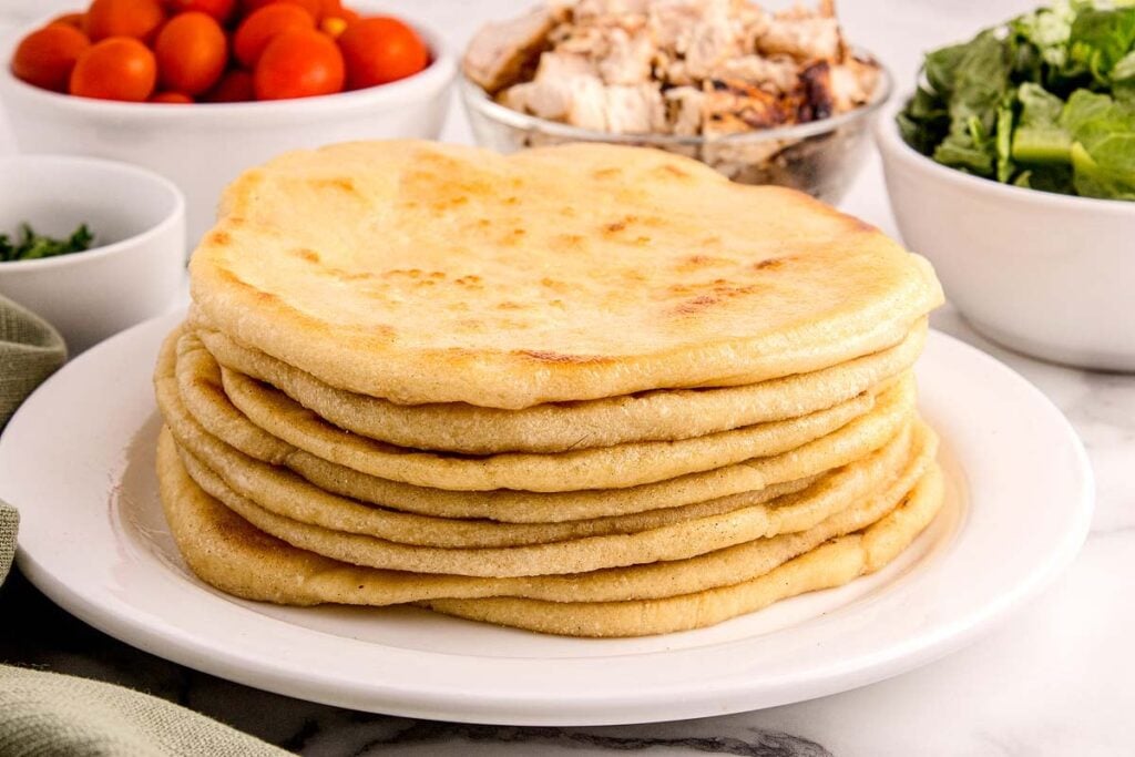 A stack of flatbreads on a white plate, with bowls of cherry tomatoes, grilled chicken, and leafy greens in the background.