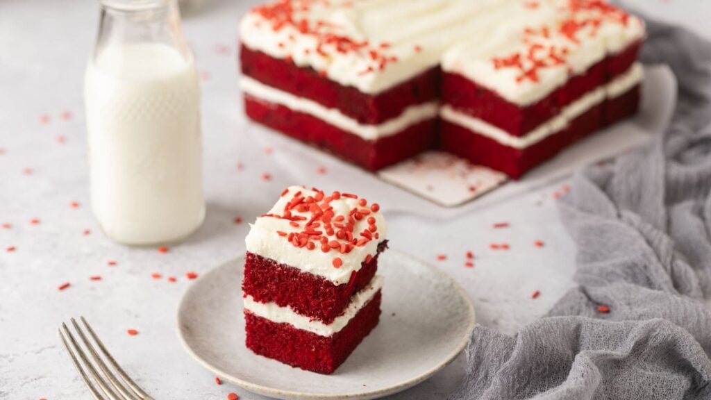 A slice of red velvet cake with white frosting and red sprinkles on a plate, next to a bottle of milk. The rest of the cake is in the background.