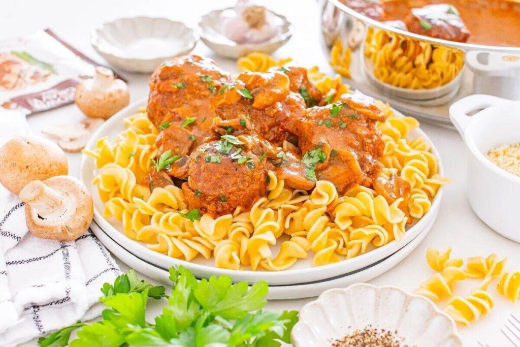 A plate of egg noodles topped with meatballs in tomato sauce, garnished with herbs, surrounded by mushrooms, parsley, and other ingredients on a white surface.
