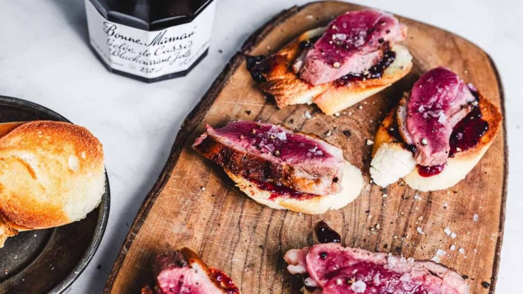 Slices of seared duck breast on toasted bread with a dark berry jam, garnished with coarse salt, served on a wooden board.