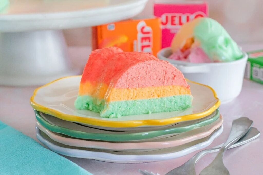 A slice of layered Jello mousse dessert in pastel colors on a stack of plates, with a bowl of ice cream and Jello boxes in the background.