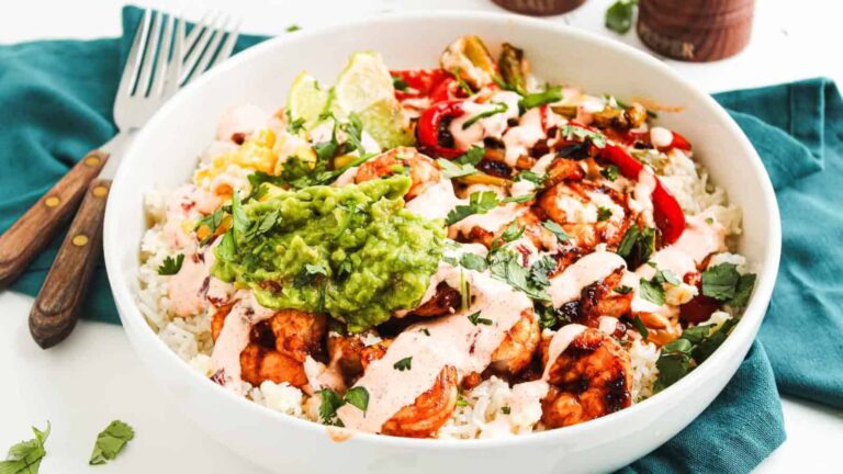 A white bowl filled with rice, grilled chicken, sautéed peppers, guacamole, lime wedges, cilantro, and drizzled with a creamy sauce, placed on a green cloth with forks nearby.