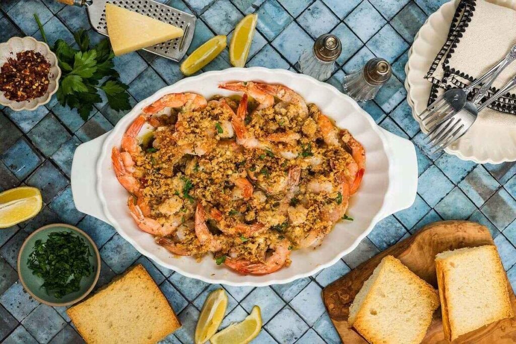 Baked shrimp with breadcrumb topping in a white dish, surrounded by lemon wedges, bread slices, grated cheese, herbs, and tableware on a blue tiled surface.