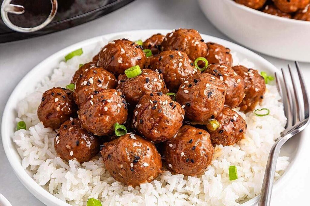 A plate of white rice topped with glazed meatballs, garnished with sliced green onions and sesame seeds, with a fork on the side.