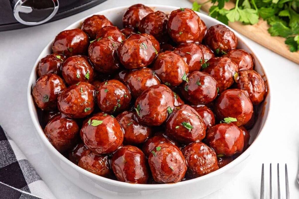 A white bowl filled with glazed meatballs garnished with chopped herbs, placed on a white surface next to a fork and parsley.