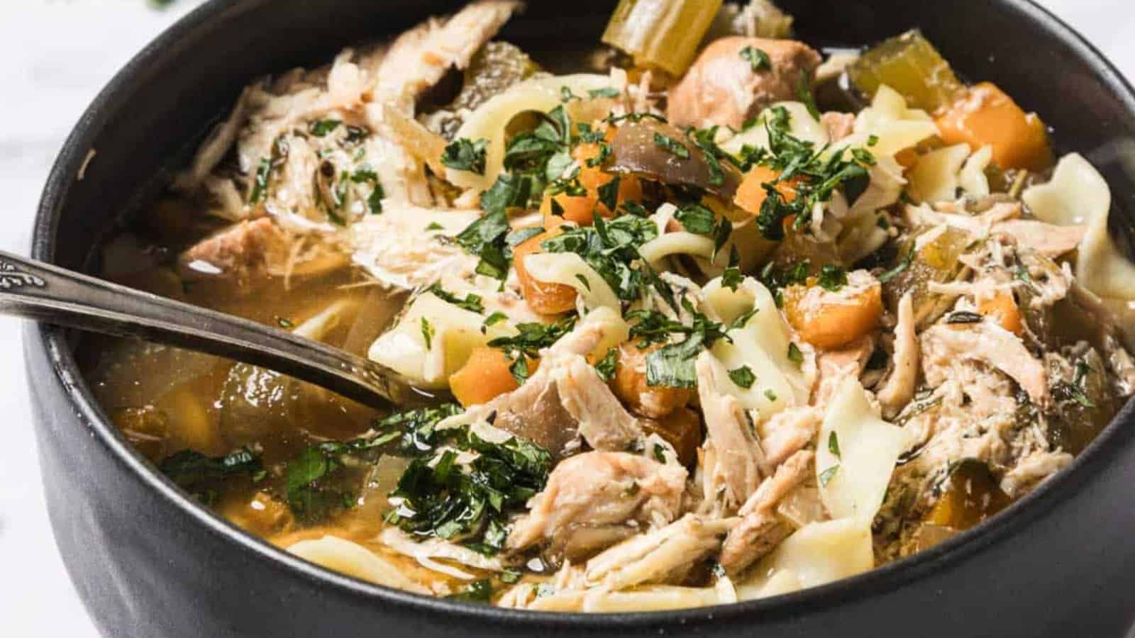 A bowl of chicken noodle soup with carrots, celery, herbs, and a spoon inside.