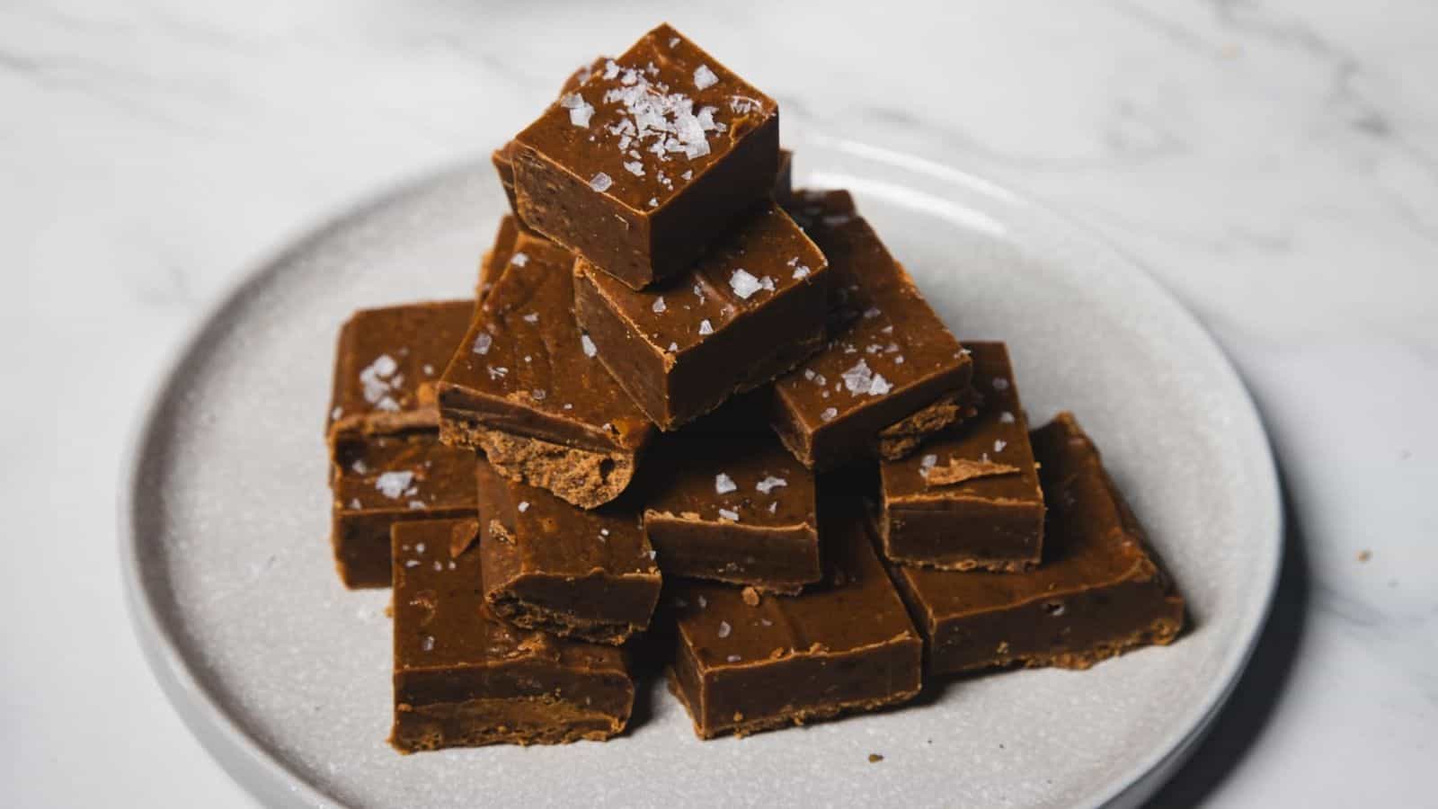 A plate of chocolate fudge squares stacked in a pyramid shape on a white surface. The fudge is topped with a sprinkle of coarse sea salt.