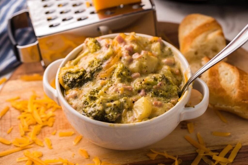 A bowl of broccoli and cheese casserole with a spoon, shredded cheese, sliced bread, and a cheese grater on a wooden board.