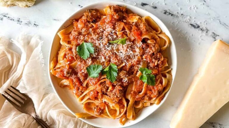 A bowl of tagliatelle pasta topped with meat sauce, grated cheese, and parsley, placed on a marble surface beside a block of cheese and a fork.