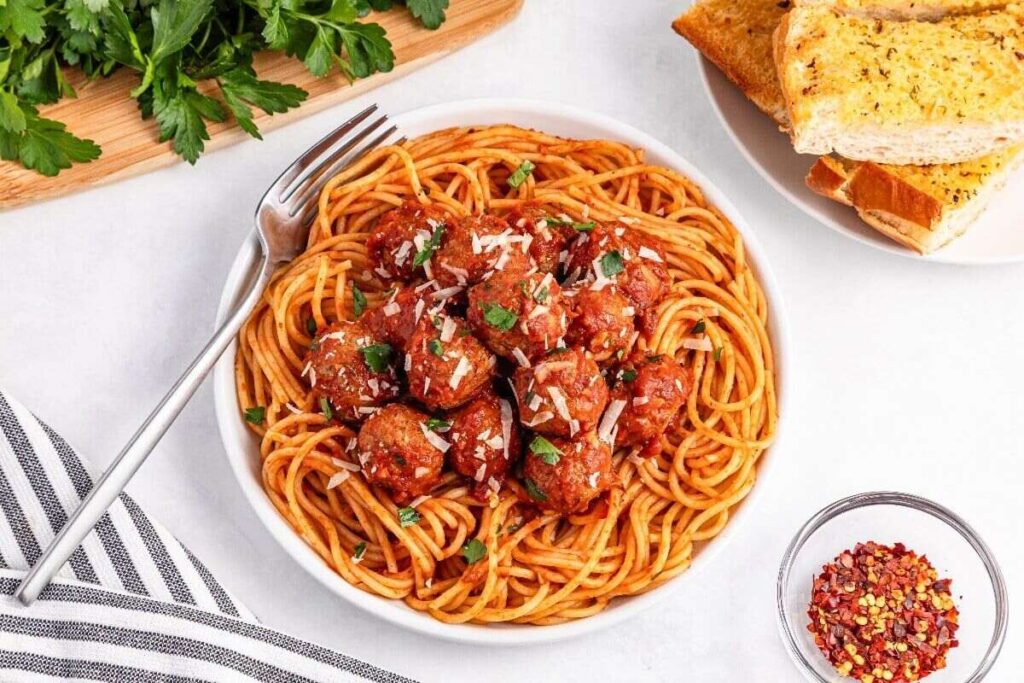 A plate of spaghetti with meatballs in tomato sauce, topped with grated cheese and parsley, served with garlic bread and a dish of red pepper flakes.