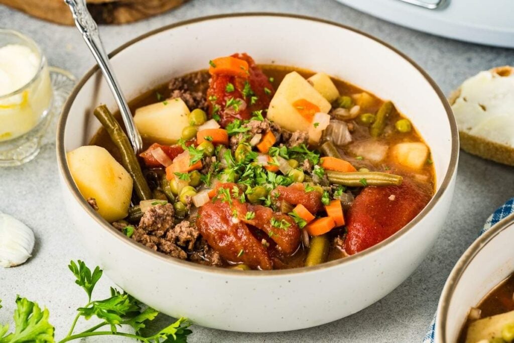 A bowl of beef and vegetable stew with potatoes, carrots, green beans, peas, and tomatoes, garnished with chopped parsley. A spoon rests in the bowl.