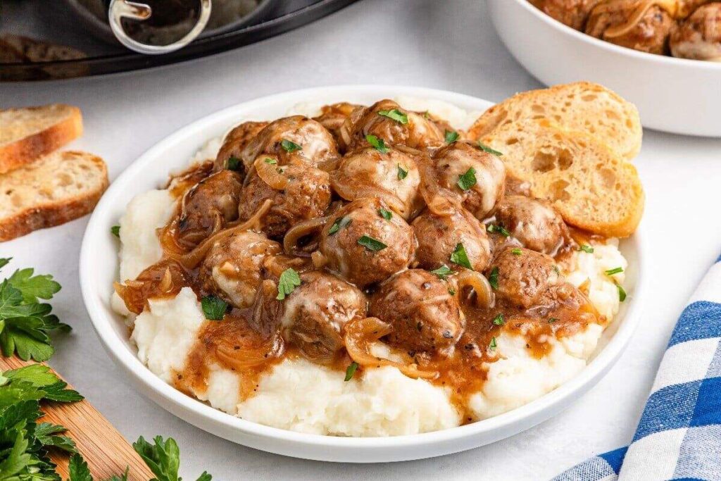 A white bowl filled with mashed potatoes topped with meatballs in brown gravy and sautéed onions, garnished with parsley, served with two slices of toasted bread.