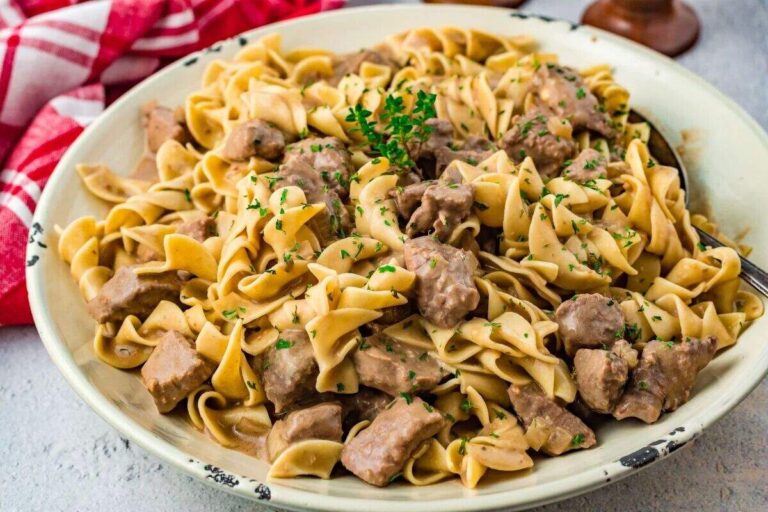 A plate of beef stroganoff with egg noodles, garnished with chopped parsley, set on a light surface with a red and white cloth in the background.