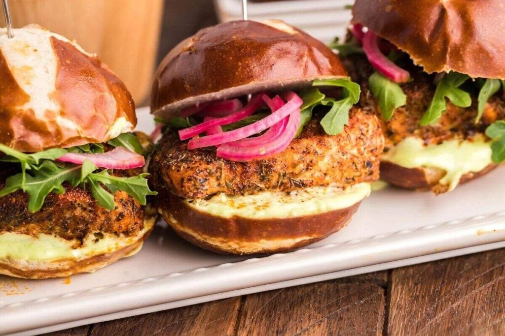 Three chicken sliders on pretzel buns with arugula, pickled onions, and Wasabi Mayo are served on a white platter.