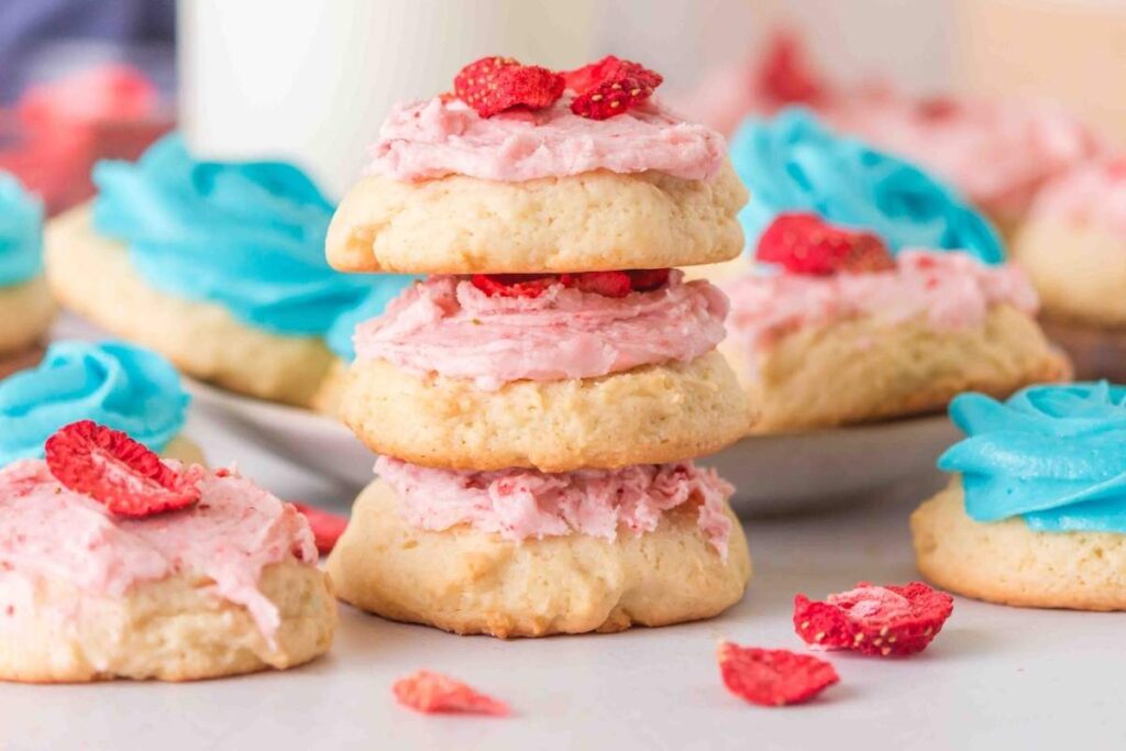 Cookies with pink and blue frosting are arranged on a plate and table, with some topped with dried strawberries and others decorated with swirls of blue icing.