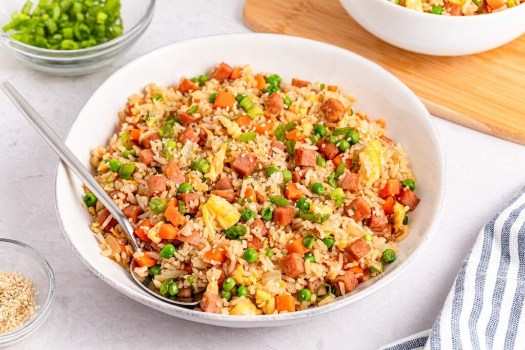 A bowl of fried rice with diced ham, peas, carrots, scrambled eggs, and a spoon. Chopped green onions and sesame seeds are in small bowls nearby.