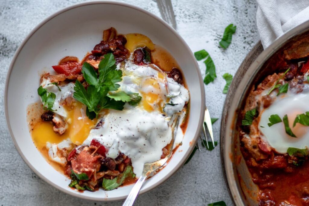 A bowl of shakshuka with poached eggs, tomato sauce, beans, fresh parsley, and a dollop of yogurt, with a fork resting inside.