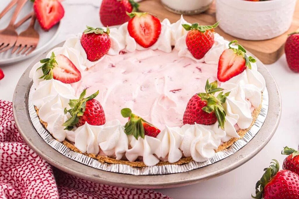 A strawberry cream pie in a foil pie pan, topped with whipped cream swirls and whole strawberries, on a white surface with extra strawberries nearby.