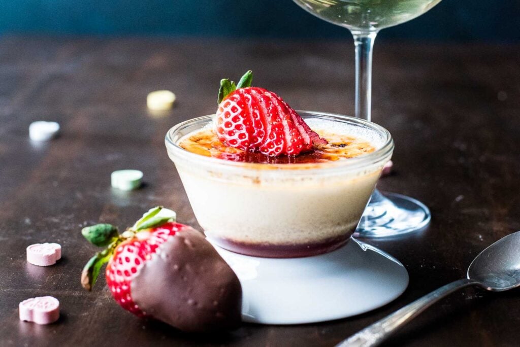 A glass dish of crème brûlée topped with sliced strawberries sits next to a chocolate-dipped strawberry, a spoon, and a wine glass on a dark surface.