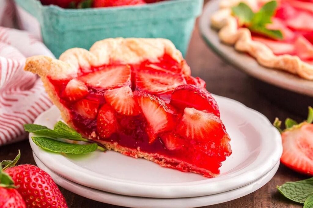 A slice of strawberry pie with a glossy filling and fresh strawberry slices sits on a white plate, garnished with a mint leaf.