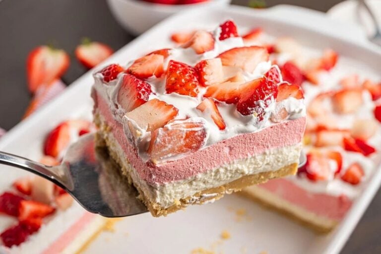 A close-up of a layered dessert with a cookie crust, cream and pink layers, topped with whipped cream and fresh strawberry pieces.