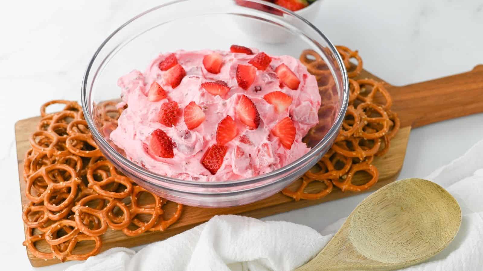 A glass bowl of pink strawberry fluff with sliced strawberries on top, surrounded by pretzels on a wooden board. A wooden spoon and white cloth are nearby.