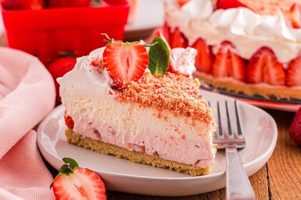 A slice of strawberry cheesecake topped with whipped cream, strawberry halves, and crumb topping on a white plate with a fork.