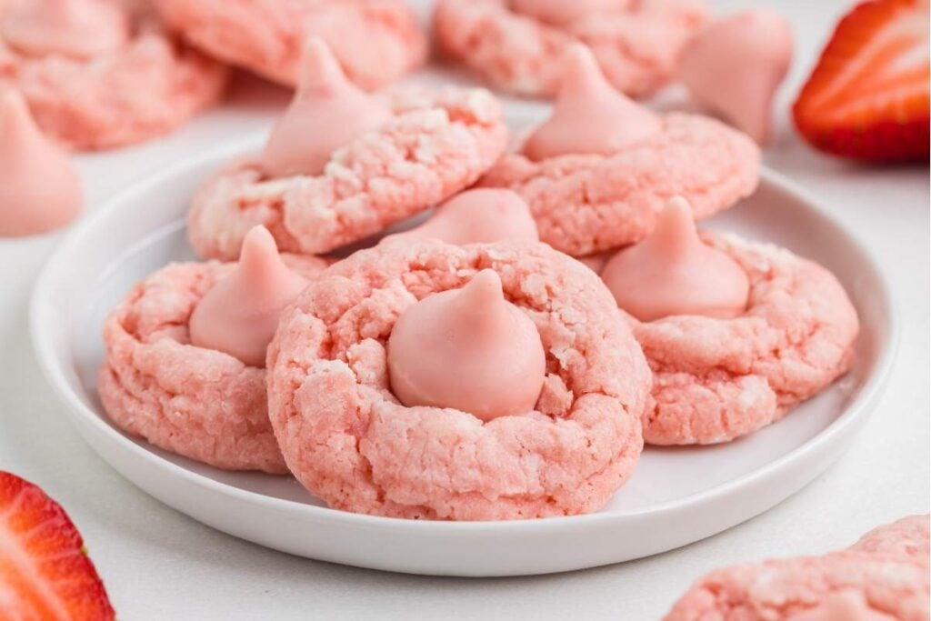 A white plate with pink strawberry cookies, each topped with a pink chocolate drop, surrounded by fresh strawberry slices.