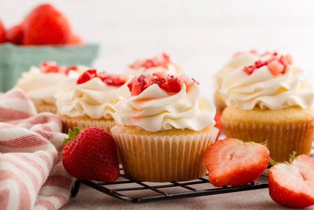 Cupcakes topped with whipped cream and chopped strawberries on a cooling rack, with whole and sliced strawberries nearby.