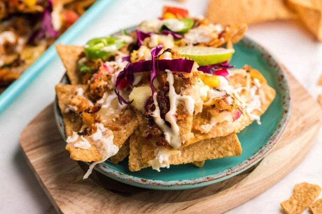 A plate of nachos topped with shredded cabbage, sliced jalapenos, lime, and drizzled with white sauce on a turquoise plate.