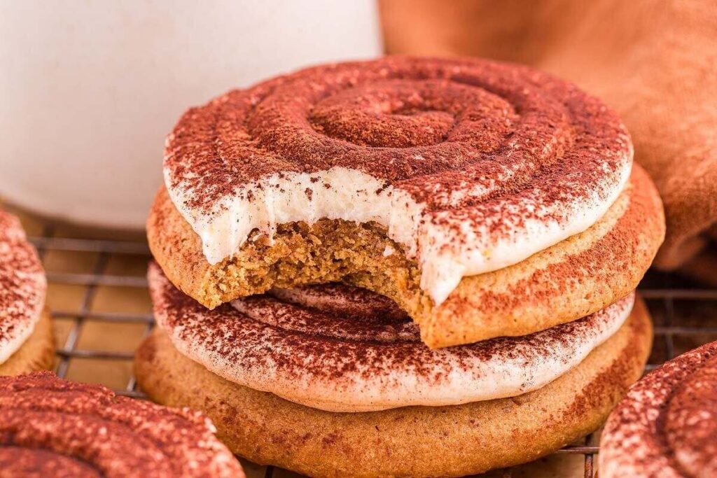 Two cookies with white icing and cocoa powder are stacked; the top cookie has a bite taken out of it. Other cookies and a mug are in the background.