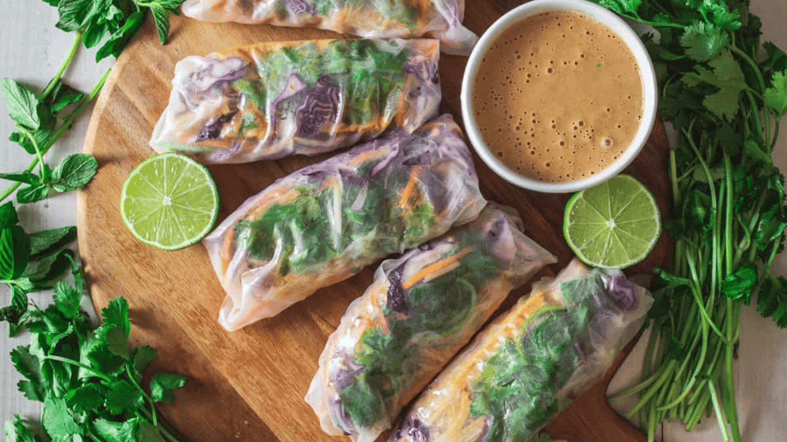 A wooden platter displays five vegetable spring rolls alongside a small bowl of brown dipping sauce. Perfect for those seeking frugal recipes, sliced limes and fresh cilantro are arranged around the platter, offering an affordable yet flavorful addition to your weekly meals.