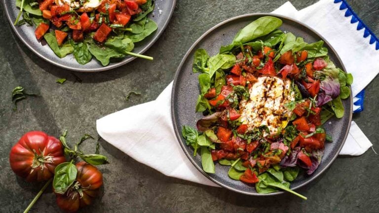 Two plates of salad with fresh spinach, tomatoes, burrata cheese, and balsamic drizzle are on a dark surface next to two heirloom tomatoes and a white napkin with a blue trim.