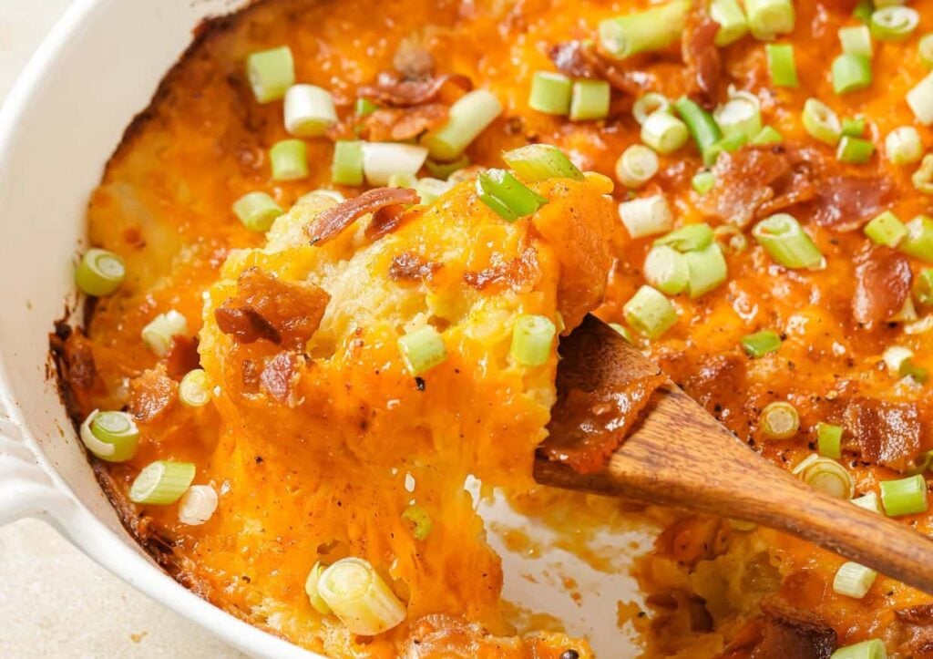A baked casserole topped with melted cheddar cheese, chopped green onions, and bacon, with a wooden spoon lifting a serving.