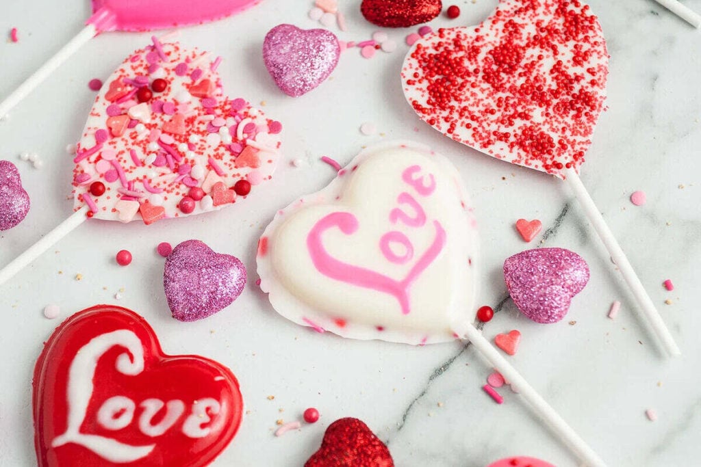 Heart-shaped lollipops with pink, red, and white decorations, some with the word "Love," surrounded by heart-shaped glitter and candy sprinkles on a marble surface.