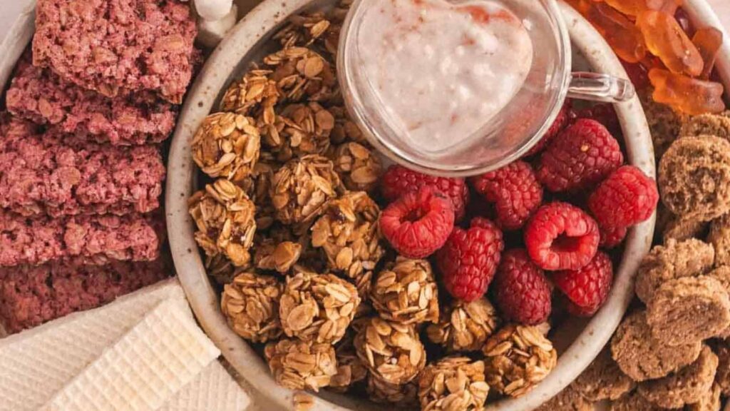 A plate with granola clusters, fresh raspberries, wafers, fruit bars, cookies, and a small cup of creamy dip.
