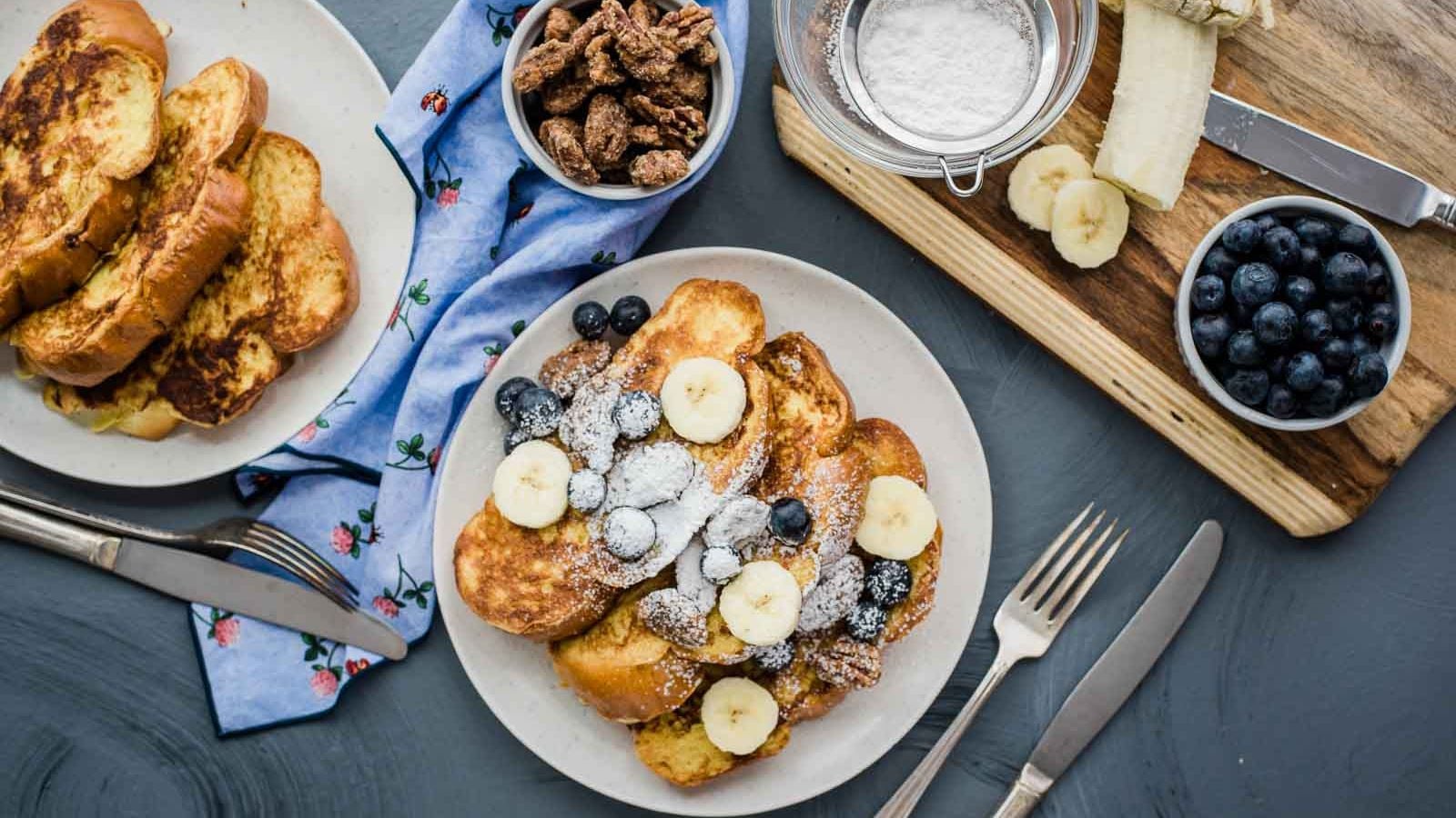 A serving of vanilla french toast next to a serving plate and bowls of candied pecans, blueberries, powdered sugar, and a banana.
